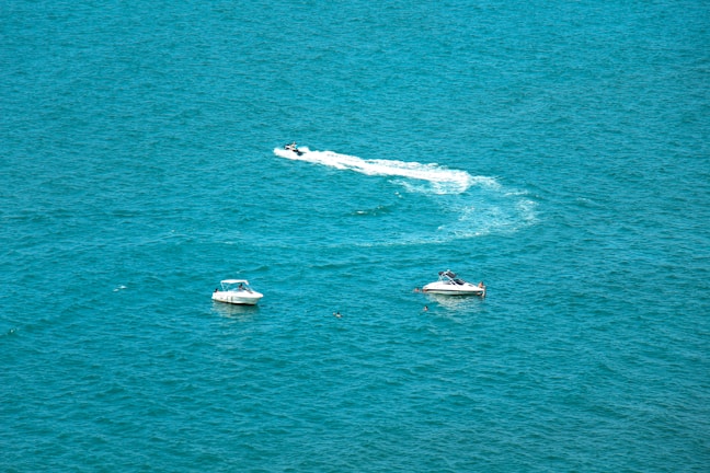 Two boats are floating on the open turquoise sea, with a jet ski creating a white wake as it speeds by, surrounded by a vast expanse of clear blue water.