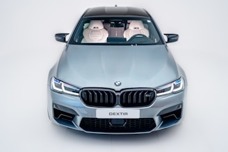 A sleek, modern car with a metallic silver finish is viewed from the front. The design features sharp headlights, a prominent grille with the BMW logo, and a license plate marked 'DEXTIR'. The interior is visible through the windshield, showing white leather seats with an embroidered logo.