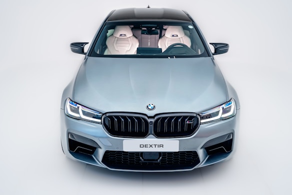 A sleek, modern car with a metallic silver finish is viewed from the front. The design features sharp headlights, a prominent grille with the BMW logo, and a license plate marked 'DEXTIR'. The interior is visible through the windshield, showing white leather seats with an embroidered logo.