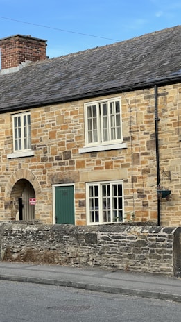 A stone cottage with a gray slate roof is featured prominently. The building has green wooden doors and multiple white-framed windows. A low stone wall runs parallel to the street in front of the house, and there is a small hanging flowerpot.