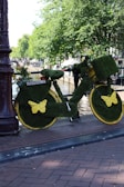 A bicycle completely covered in green artificial grass is parked by the side of a canal. The wheels have yellow butterfly decorations, and there are small pot plants on the carrier. The area is surrounded by lush green trees, and a waterway is visible nearby. The setting suggests a lively urban environment.