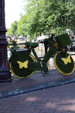 A bicycle completely covered in green artificial grass is parked by the side of a canal. The wheels have yellow butterfly decorations, and there are small pot plants on the carrier. The area is surrounded by lush green trees, and a waterway is visible nearby. The setting suggests a lively urban environment.