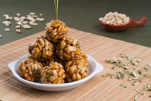 A white dish contains a stack of sweet treats made from peanuts and sugar syrup, forming round clusters. The background features scattered peanuts and dried leaves on a woven mat, with a small bowl of peanuts to the side. The image captures a drizzle of syrup being poured over the top treat, enhancing its glossy appearance.