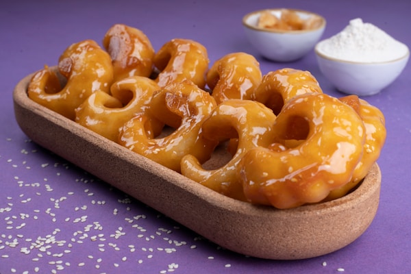 Glazed, twisted donuts are artfully arranged in an oval wooden tray against a purple background. Two small bowls, one containing flour and the other holding a sauce or dip, are visible in the background. Sesame seeds are sprinkled across the surface.