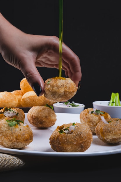 A hand holds a piece of crispy, round Indian snack above a white plate. Other similar snacks are arranged on the plate. Small bowls with dips and garnishes are in the background. The snacks are garnished with chopped onions and herbs.