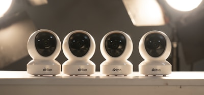 Four white security cameras are lined up on a surface with soft studio lighting in the background. Each camera is identical in design, with a spherical shape and a black lens visible in the front. The lighting creates a muted, professional atmosphere.