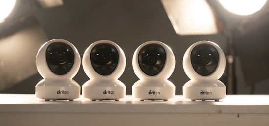 Four white security cameras are lined up on a surface with soft studio lighting in the background. Each camera is identical in design, with a spherical shape and a black lens visible in the front. The lighting creates a muted, professional atmosphere.
