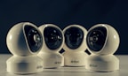 Four spherical security cameras with a glossy white finish are positioned in a row, each featuring a prominent lens and the brand name 'Briton' visible on their bases. The background is dark, contrasting with the brightly lit cameras.