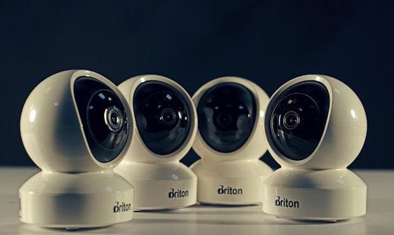 Four spherical security cameras with a glossy white finish are positioned in a row, each featuring a prominent lens and the brand name 'Briton' visible on their bases. The background is dark, contrasting with the brightly lit cameras.