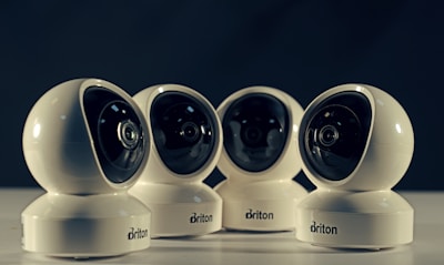 Four spherical security cameras with a glossy white finish are positioned in a row, each featuring a prominent lens and the brand name 'Briton' visible on their bases. The background is dark, contrasting with the brightly lit cameras.
