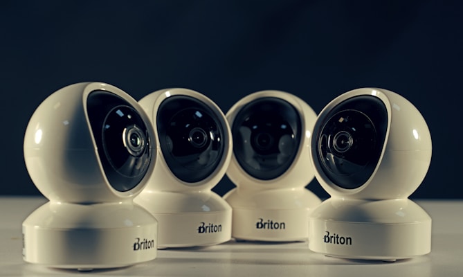 Four spherical security cameras with a glossy white finish are positioned in a row, each featuring a prominent lens and the brand name 'Briton' visible on their bases. The background is dark, contrasting with the brightly lit cameras.