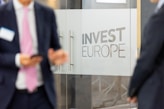 Two individuals in business attire are standing near glass doors with the phrase 'Invest Europe' etched on them. One person is holding a smartphone.