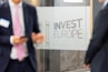 Two individuals in business attire are standing near glass doors with the phrase 'Invest Europe' etched on them. One person is holding a smartphone.