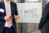 Two individuals in business attire are standing near glass doors with the phrase 'Invest Europe' etched on them. One person is holding a smartphone.
