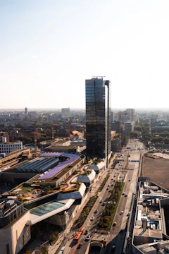 A modern urban scene featuring a tall glass skyscraper surrounded by various buildings and roads. The urban landscape includes a railway track with a bridge and several vehicles on the roads, emphasizing the city's infrastructure. The surrounding architecture is diverse, with a mix of contemporary and functional designs, set against a clear sky.