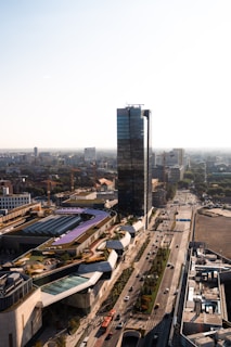 A modern urban scene featuring a tall glass skyscraper surrounded by various buildings and roads. The urban landscape includes a railway track with a bridge and several vehicles on the roads, emphasizing the city's infrastructure. The surrounding architecture is diverse, with a mix of contemporary and functional designs, set against a clear sky.