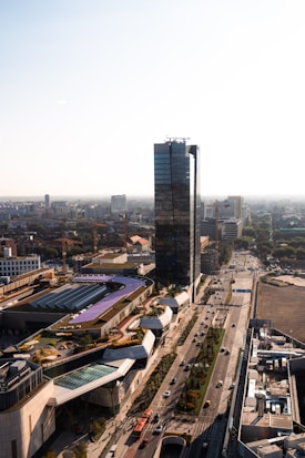 A modern urban scene featuring a tall glass skyscraper surrounded by various buildings and roads. The urban landscape includes a railway track with a bridge and several vehicles on the roads, emphasizing the city's infrastructure. The surrounding architecture is diverse, with a mix of contemporary and functional designs, set against a clear sky.