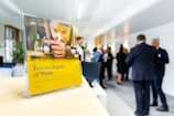 A book titled 'Private Equity at Work' is positioned prominently on a wooden surface in the foreground. In the background, a group of people in business attire are engaged in conversation, suggesting a professional setting such as a conference or networking event.