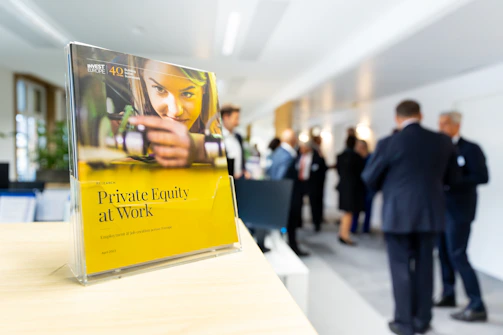 A book titled 'Private Equity at Work' is positioned prominently on a wooden surface in the foreground. In the background, a group of people in business attire are engaged in conversation, suggesting a professional setting such as a conference or networking event.