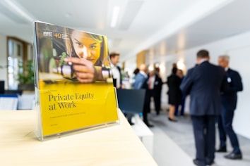 A book titled 'Private Equity at Work' is positioned prominently on a wooden surface in the foreground. In the background, a group of people in business attire are engaged in conversation, suggesting a professional setting such as a conference or networking event.