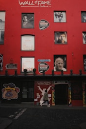 A vibrant red wall features multiple photographs of famous musicians and a sign for the Irish Rock 'n' Roll Museum Experience. A person in a hoodie is walking along the cobblestone street in front of the wall. The words 'Wall of Fame Dublin' are prominently painted at the top.