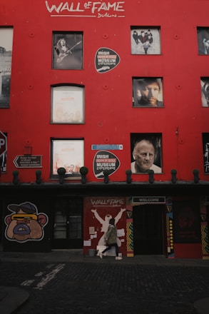 A vibrant red wall features multiple photographs of famous musicians and a sign for the Irish Rock 'n' Roll Museum Experience. A person in a hoodie is walking along the cobblestone street in front of the wall. The words 'Wall of Fame Dublin' are prominently painted at the top.