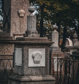 A stone monument in a cemetery features a sculpted face with simple geometric patterns. The design includes several stone pillars adorned with urn-shaped ornaments, and cast iron fences surround the structures. The scene is set in a shaded area with trees in the background, suggesting a serene yet somber atmosphere.