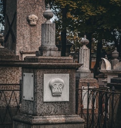 A stone monument in a cemetery features a sculpted face with simple geometric patterns. The design includes several stone pillars adorned with urn-shaped ornaments, and cast iron fences surround the structures. The scene is set in a shaded area with trees in the background, suggesting a serene yet somber atmosphere.