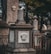 A stone monument in a cemetery features a sculpted face with simple geometric patterns. The design includes several stone pillars adorned with urn-shaped ornaments, and cast iron fences surround the structures. The scene is set in a shaded area with trees in the background, suggesting a serene yet somber atmosphere.