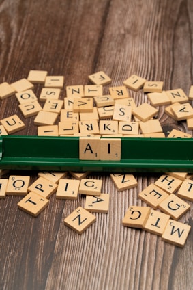 A collection of wooden letter tiles with black letters scattered across a wooden surface. Three tiles are neatly arranged on a green tile rack, spelling out the letters 'A', 'I'. The surrounding tiles are in a random arrangement.