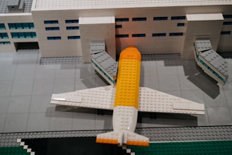A detailed Lego model of an airport terminal with a plane parked at the gate. The building is made of white Lego bricks with blue windows, and the plane has a distinctive yellow and white color scheme. The scene includes jet bridges connecting the plane to the terminal.