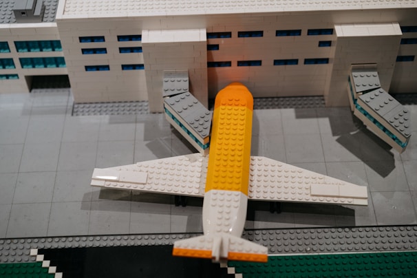 A detailed Lego model of an airport terminal with a plane parked at the gate. The building is made of white Lego bricks with blue windows, and the plane has a distinctive yellow and white color scheme. The scene includes jet bridges connecting the plane to the terminal.