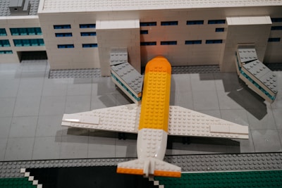 A detailed Lego model of an airport terminal with a plane parked at the gate. The building is made of white Lego bricks with blue windows, and the plane has a distinctive yellow and white color scheme. The scene includes jet bridges connecting the plane to the terminal.