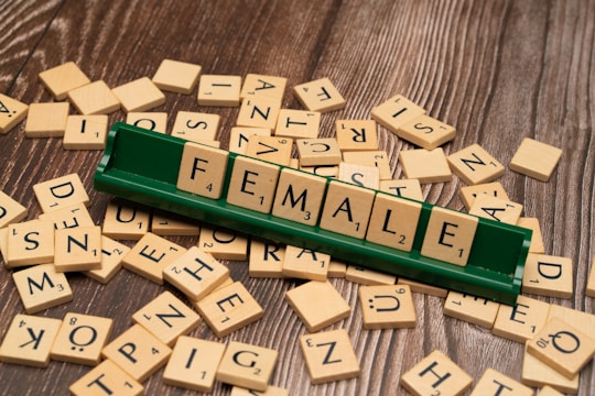 Wooden alphabet tiles are scattered on a wooden surface with a green tile holder displaying the word 'FEMALE' using the tiles.