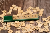 Wooden letter tiles scattered across a wooden surface, with a green tile holder displaying the word 'PODCAST' spelled out prominently.