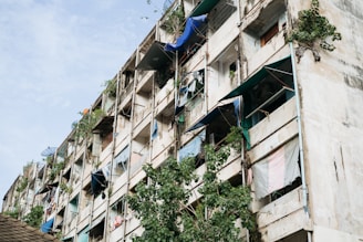 A large, weathered apartment building with multiple floors and visible signs of neglect. Exposed balconies show mismatched tarps, sheets, and makeshift covers. Plants grow sporadically on the structure, adding to the worn appearance. The building's facade is peeling, with visible cracks and patches, giving it a rugged look.
