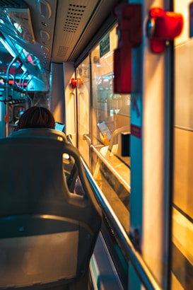 An interior view of a public bus showing a row of seats and the passageway. A person is seated, facing forward, and looking at a smartphone. The bus interior has a modern design with overhead lights and handrails.