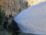 A powerful jet of water is being released from a structure built into the side of a steep, rocky cliff. Surrounding the water, lush greenery and moss are present, contrasting with the dry, craggy rock formation. The water is cascading with great force, creating a dramatic spray effect.