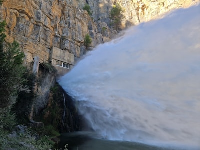 A powerful jet of water is being released from a structure built into the side of a steep, rocky cliff. Surrounding the water, lush greenery and moss are present, contrasting with the dry, craggy rock formation. The water is cascading with great force, creating a dramatic spray effect.