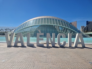 Large architectural letters spelling out 'VALÈNCIA' are prominently displayed in front of a modern, dome-shaped glass building. The structure features a futuristic design with a reflective surface and is situated next to a body of water. In the background, a clear blue sky complements tall city buildings.