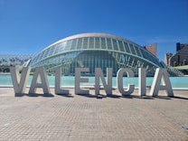 Large architectural letters spelling out 'VAL&Egrave;NCIA' are prominently displayed in front of a modern, dome-shaped glass building. The structure features a futuristic design with a reflective surface and is situated next to a body of water. In the background, a clear blue sky complements tall city buildings.