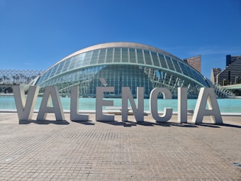 Large architectural letters spelling out 'VAL&Egrave;NCIA' are prominently displayed in front of a modern, dome-shaped glass building. The structure features a futuristic design with a reflective surface and is situated next to a body of water. In the background, a clear blue sky complements tall city buildings.