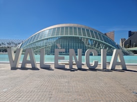 Large architectural letters spelling out 'VAL&Egrave;NCIA' are prominently displayed in front of a modern, dome-shaped glass building. The structure features a futuristic design with a reflective surface and is situated next to a body of water. In the background, a clear blue sky complements tall city buildings.