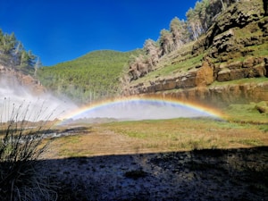 A vibrant rainbow arcs across a natural landscape, surrounded by lush greenery and rocky cliffs. The foreground features a misty spray that adds a mystical quality to the scene, with a bright blue sky overhead.