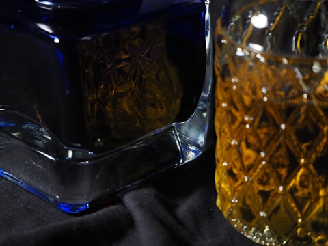 A close-up of two glass containers with intricate textured surfaces. The object on the left appears to be a square-shaped decanter in a rich blue hue, while the one on the right is a tumbler with a diamond pattern and a warm brown liquid inside. They are placed on a dark fabric background.