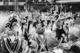 A large gathering of people dressed in traditional attire with intricate patterns and headdresses featuring feathers. The people appear to be part of a ceremonial or cultural event with a sense of movement and unity in their arrangement.