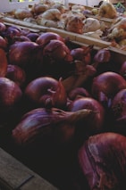 A collection of onions is displayed in a market setting. The foreground features red onions with their deep purplish skins, while the background shows lighter colored onions in crates. Natural light illuminates the scene, enhancing the texture and layers of the onion skins.