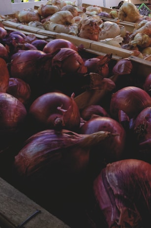 A collection of onions is displayed in a market setting. The foreground features red onions with their deep purplish skins, while the background shows lighter colored onions in crates. Natural light illuminates the scene, enhancing the texture and layers of the onion skins.