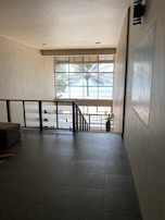 A modern interior space with a large window revealing a view of a palm tree and the ocean outside. The room has tiled flooring and neutral-colored walls, with a staircase leading downwards. A potted plant is near the staircase, adding a touch of greenery to the minimalist design.