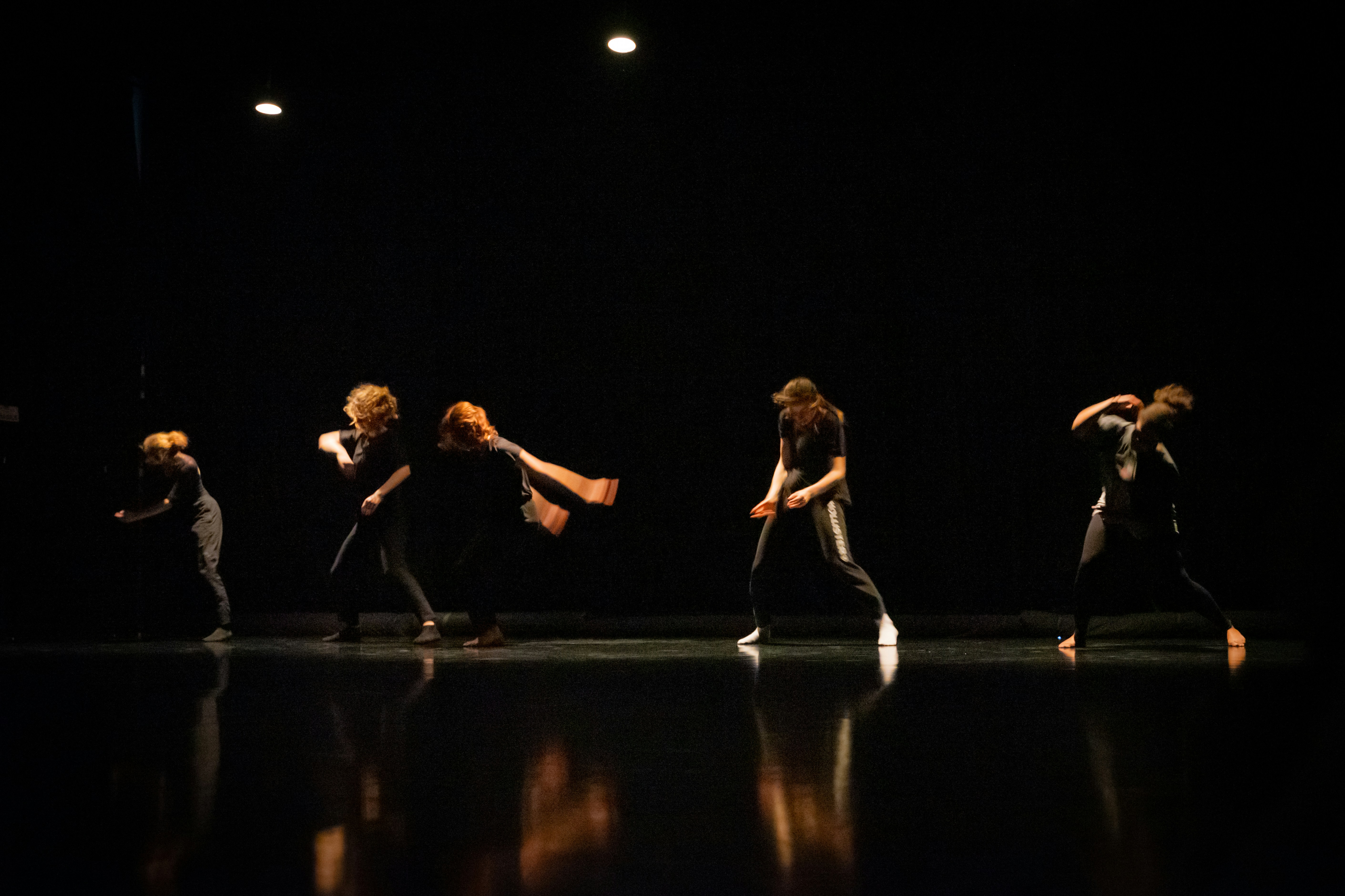A group of dancers performing on a dimly lit stage, their movements captured in mid-action with dramatic lighting highlighting their forms against the dark background.